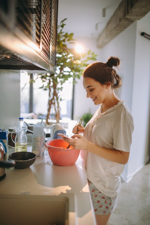 A young woman makes pancakes in her kitchen while watching a virtual breakfast class on her phone.