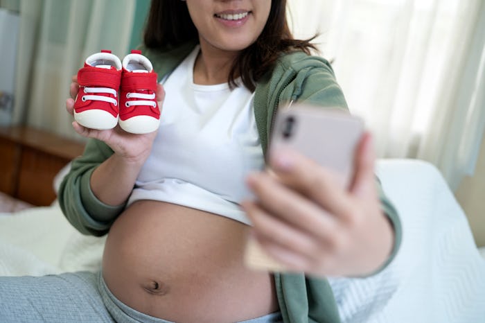 Pregnant woman taking selfie with baby shoes in bedroom at home.