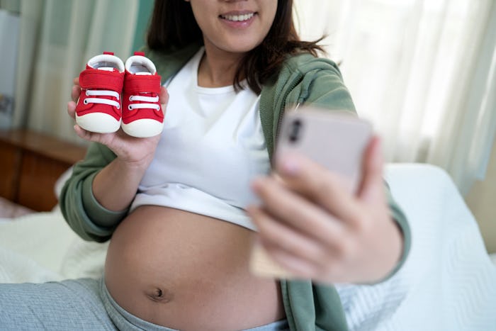 Pregnant woman taking selfie with baby shoes in bedroom at home.