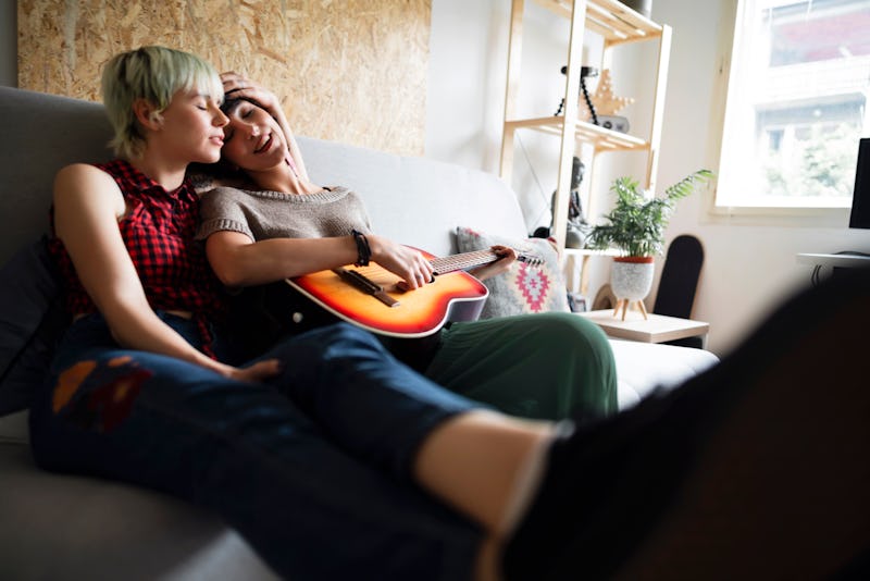 Two young Caucasian women in love sitting on the sofa at home. Spending their time together and relaxing while playing the guitar.