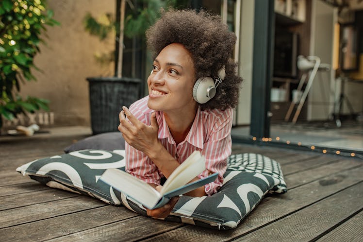 Mixed race woman is relaxing at the home balcony, laying down on the pillows and reading a book