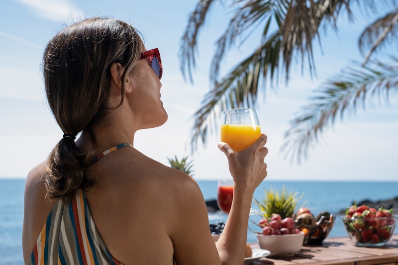 Woman sipping juice overlooking the beach. Caucasian woman enjoying breakfast in the morning by the ...