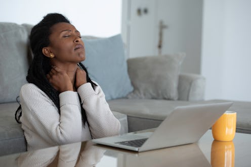 A person massages their own neck while closing their eyes and looking away from their laptop. Fatigu...