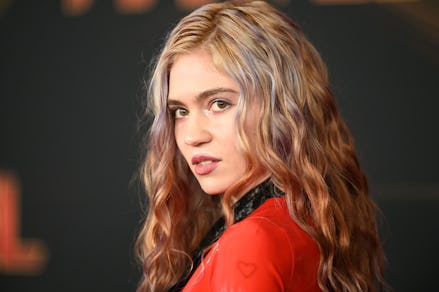 Grimes in a red and black blazer, with a wavy hairstyle, looking to the side
