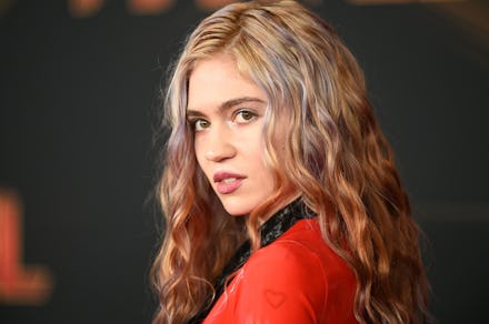 Grimes in a red and black blazer, with a wavy hairstyle, looking to the side