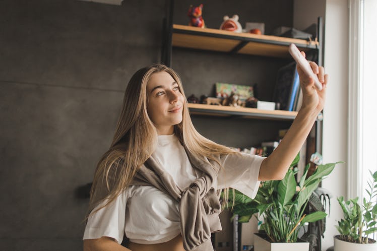 A glowing woman takes a selfie at home, showing off her hair parted in the middle.