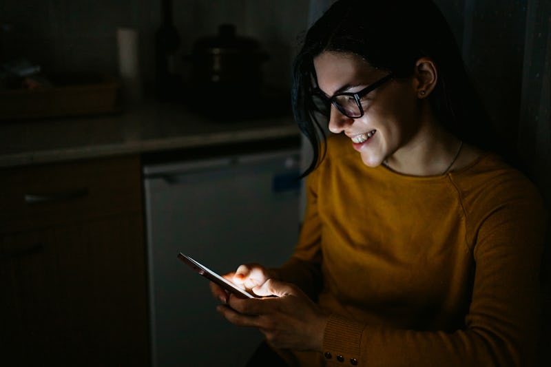 Young Caucasian pretty woman using mobile phone in home kitchen at night.