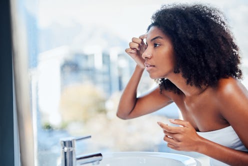 A person with natural hair wears a white towel in the bathroom while checking their face in the mirr...