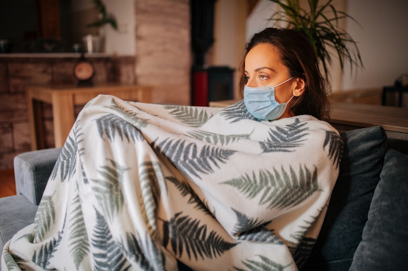 A person sits on their couch, covered in a blanket while wearing a mask. If your friend has COVID, t...
