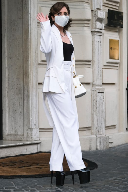Lady Gaga in a black top and a white suit, with a white face mask and black fetish heels waving
