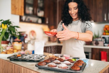 A smiling woman coats muffins in powdered sugar in her kitchen.