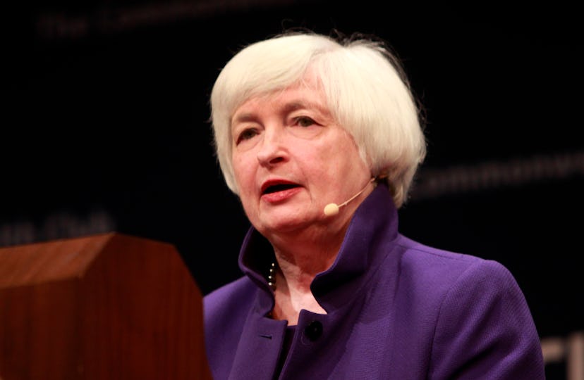Dr. Janet Yellen wears a purple shirt and stands behind a podium. 