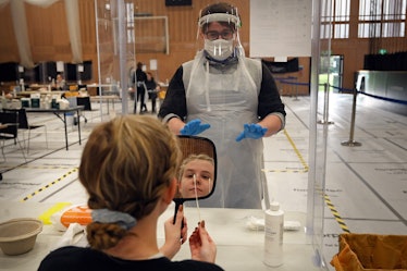 A medical worker in protective gear helps a young woman holding a mirror with a COVID-19 test