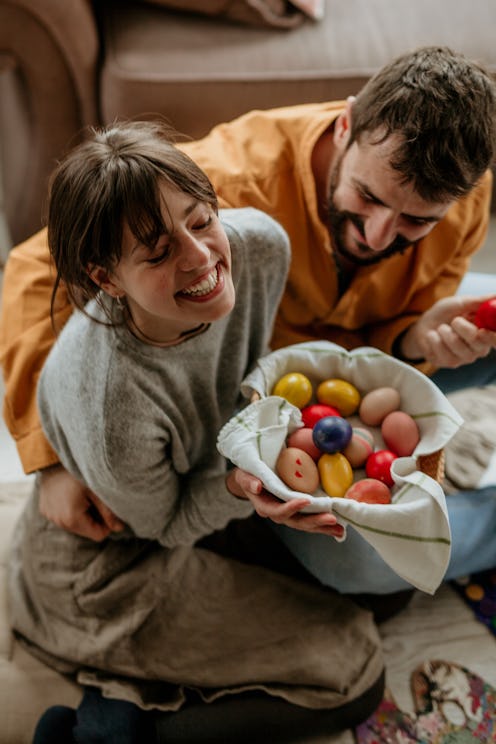 Top view of a loving couple painting Easter eggs in a cozy living room, crate full of colorful eggs ...