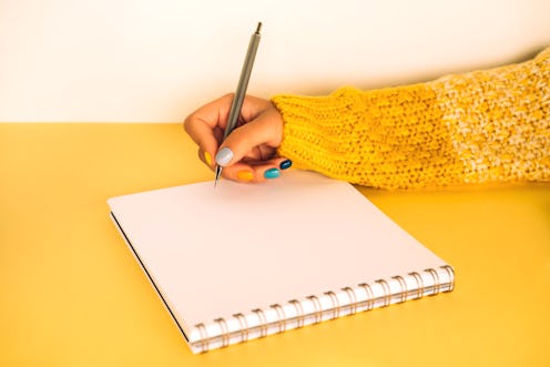 Woman in yellow white knitted sweater with bright multi colored manicure is writing in notebook sitt...
