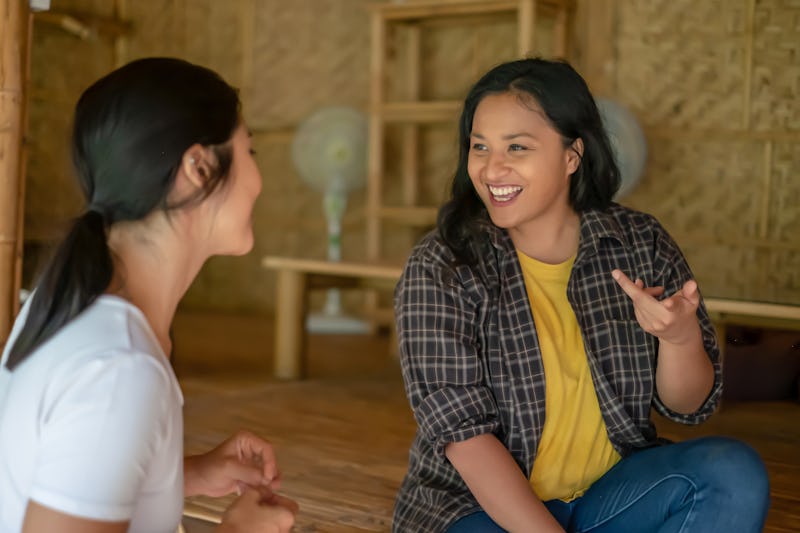 Candid shot of two cheerful Malaysian volunteers chatting on the porch of a sustainable bamboo build...