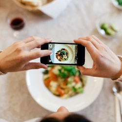 Bird's eye view of a woman taking a photo of her dinner with her smartphone. The plate is seen throu...