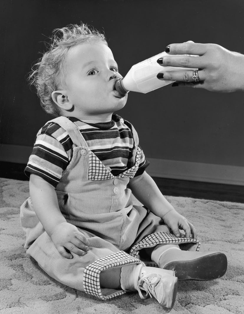 circa 1945:  Full-length image of an infant sitting a on a carpet and drinking from a bottle held by...