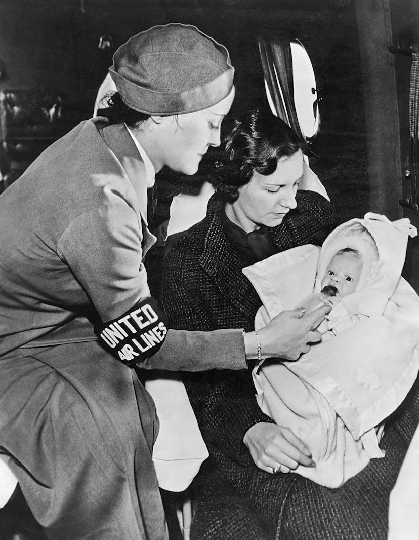 A United Airlines stewardess gives a baby a bottle on the airplane while the mother looks on, mid to...