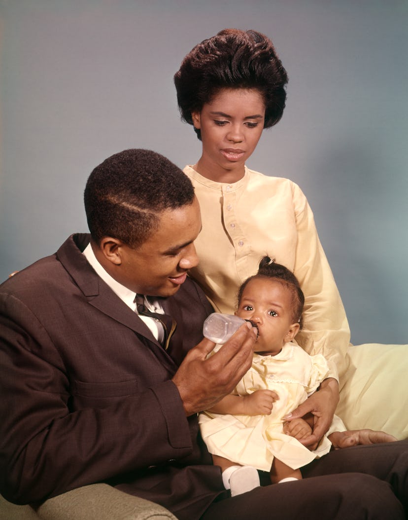 1960s AFRICAN AMERICAN FAMILY FATHER FEEDING BABY BOTTLE MOTHER SITTING ON ARM OF CHAIR  (Photo by H...