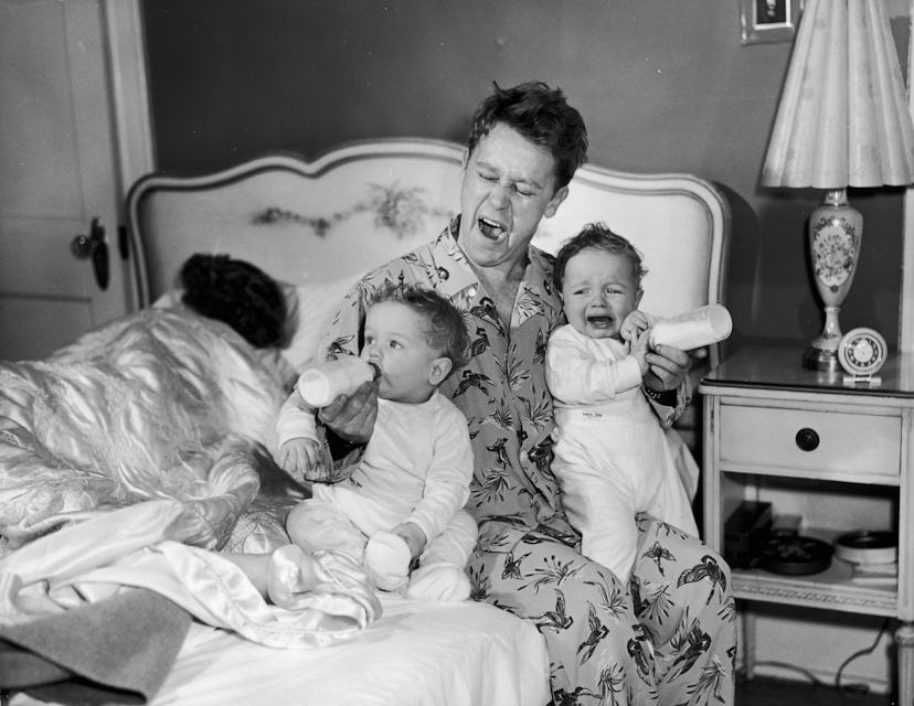 circa 1955:  A father sits on the edge of his bed, yawning as he feeds twin babies bottles of milk. ...