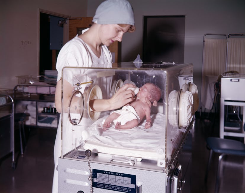 1960s WOMAN NURSE BOTTLE FEEDING INFANT BABY GIRL IN HOSPITAL NURSERY ISOLETTE PREMATURE INTENSIVE C...