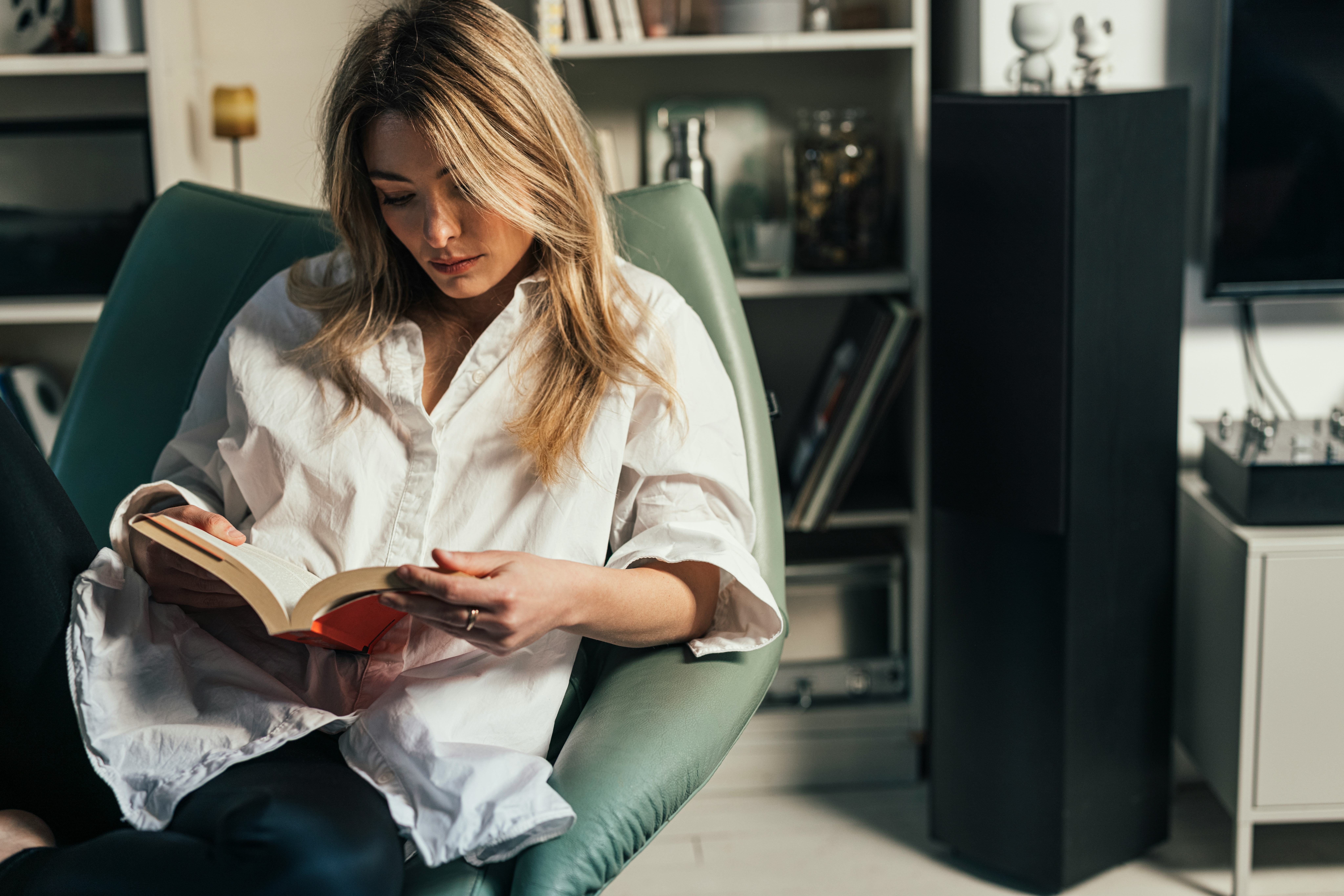 Blond elegant woman in a white shirt sitting casually in her library, reading her favorite poetry bo...
