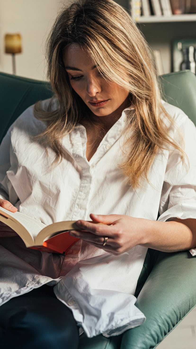 Blond elegant woman in a white shirt sitting casually in her library, reading her favorite poetry bo...