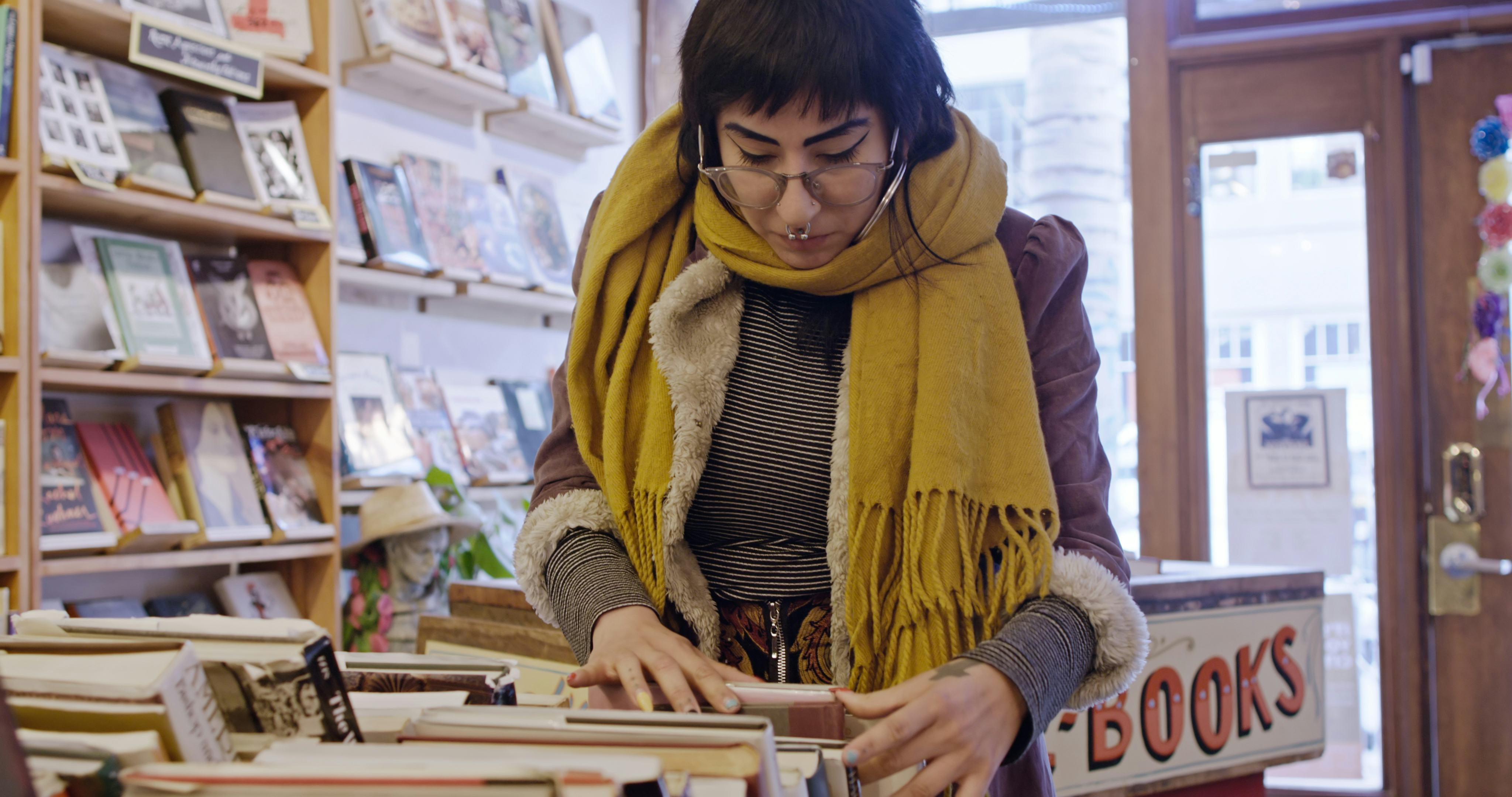 A young Latina woman with piercings and tattoos browsing through a box of books in a used bookstore.