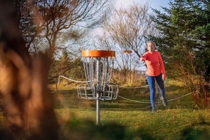 Woman tossing a disc into the basket goal. Caucasian woman playing disc golf game outdoors in a park...