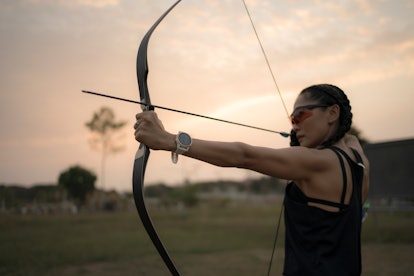 Person holding a bow and arrow at the ready, outdoors at sunset. 