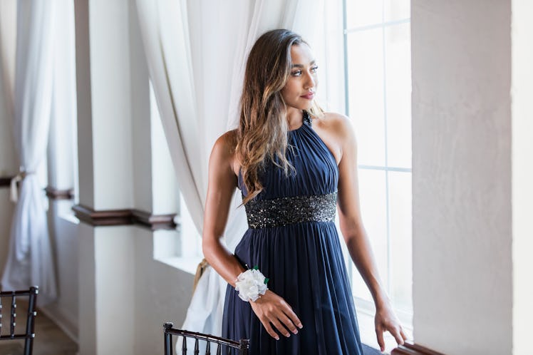A girl stands next to a window wearing her prom dress.