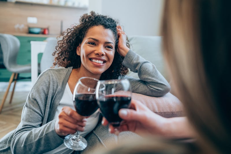 Shot of two happy women sitting on the floor making small talk and drinking wine. Smiling friends to...