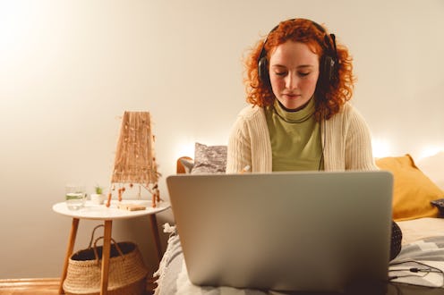 Copy space shot of young redhead sitting on her bed, listening to music via headphones and typing ho...