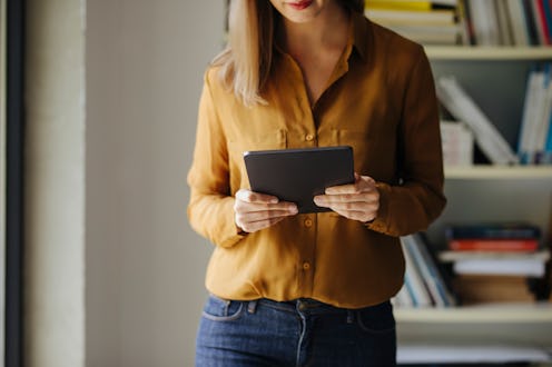 An unrecognizable businesswoman standing in her office by the window and reading on her tablet