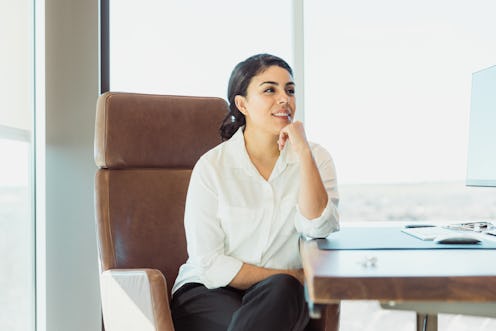 The mid adult female entrepreneur takes a break from work to daydream.  She is sitting at her desk i...