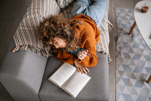 Young pretty student woman lying down on the sofa, reading book, studying for exam.