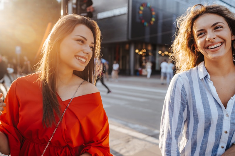 Two young cheerful female friends walking and smiling in the city on sunny day