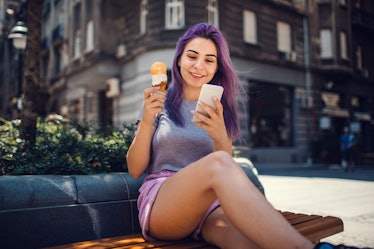 Young woman with purple hairs eating ice cream in the city and using smart phone.