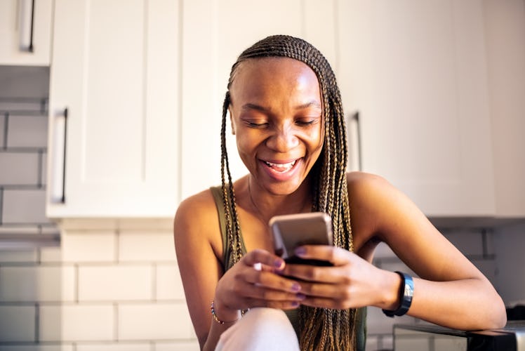 Happy young African woman sitting in her kitchen while texting on smart phone and smiling
