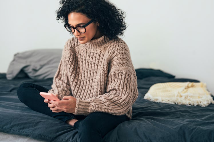 Beautiful young woman in knitwear sitting on her bed and texting on her smartphone (copy space)
