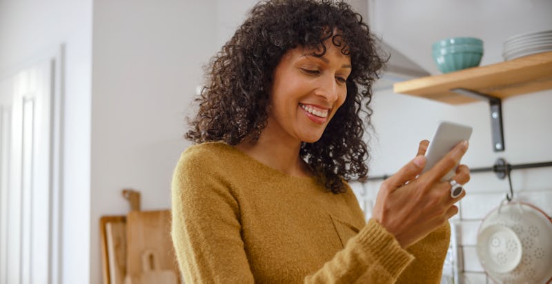 Happy young woman using her smartphone while standing in kitchen. Here's how to hide your likes on Instagram.