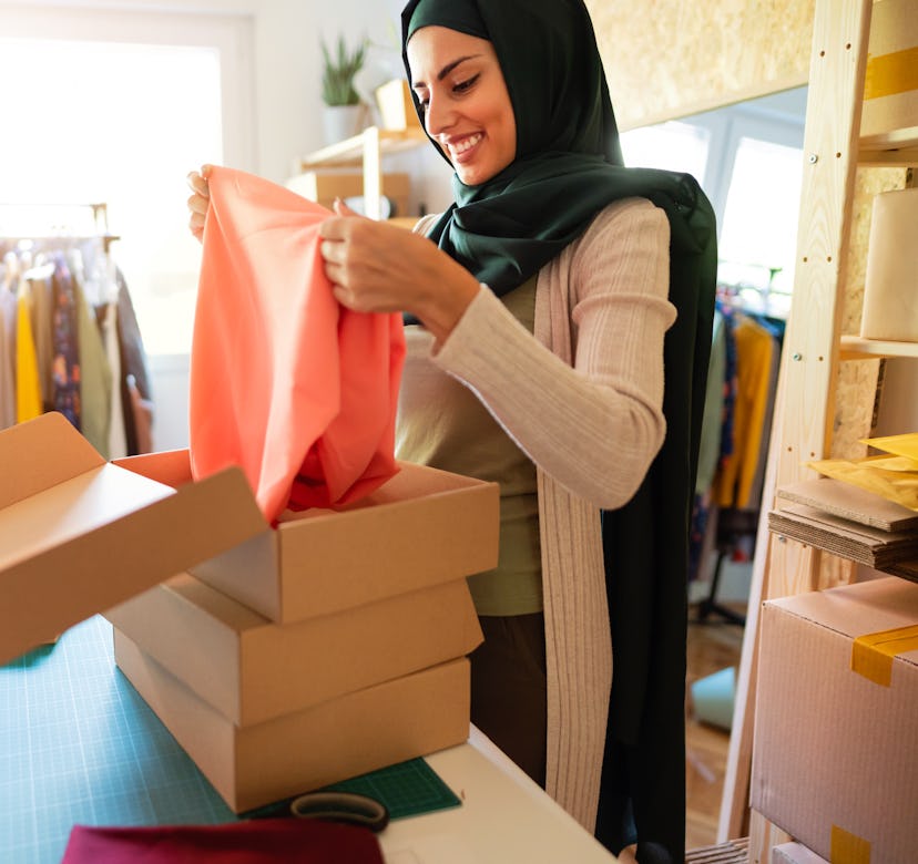 Smiling Muslim woman with hijab packing product into a cardboard box and preparing it for a delivery...