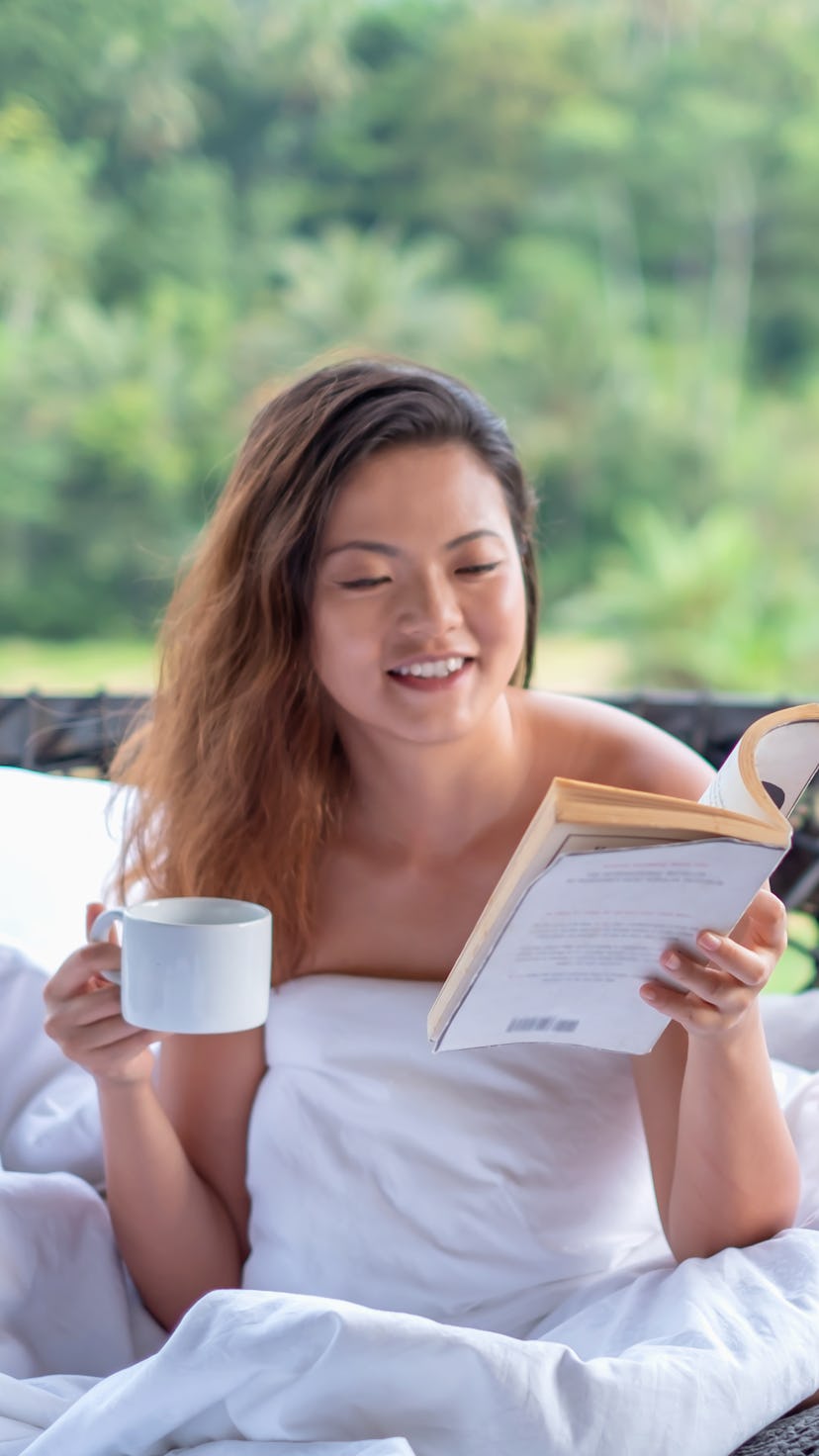 Shot of a beautiful Asian traveler reading book while drinking her morning coffee. She's sitting on ...