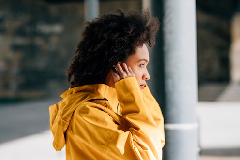 A portrait of a beautiful young woman in a yellow jacket, putting earphones on and preparing for a j...
