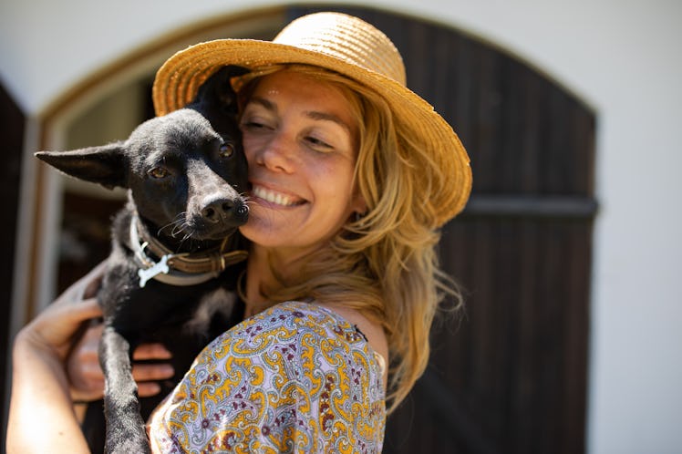 A pretty young woman with a hat is holding a cute black mixed breed in her arms and smiling