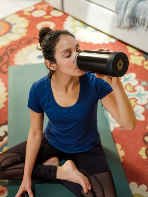 A young latin woman wearing sports clothes at home and drinking water from a reusable bottle after a...