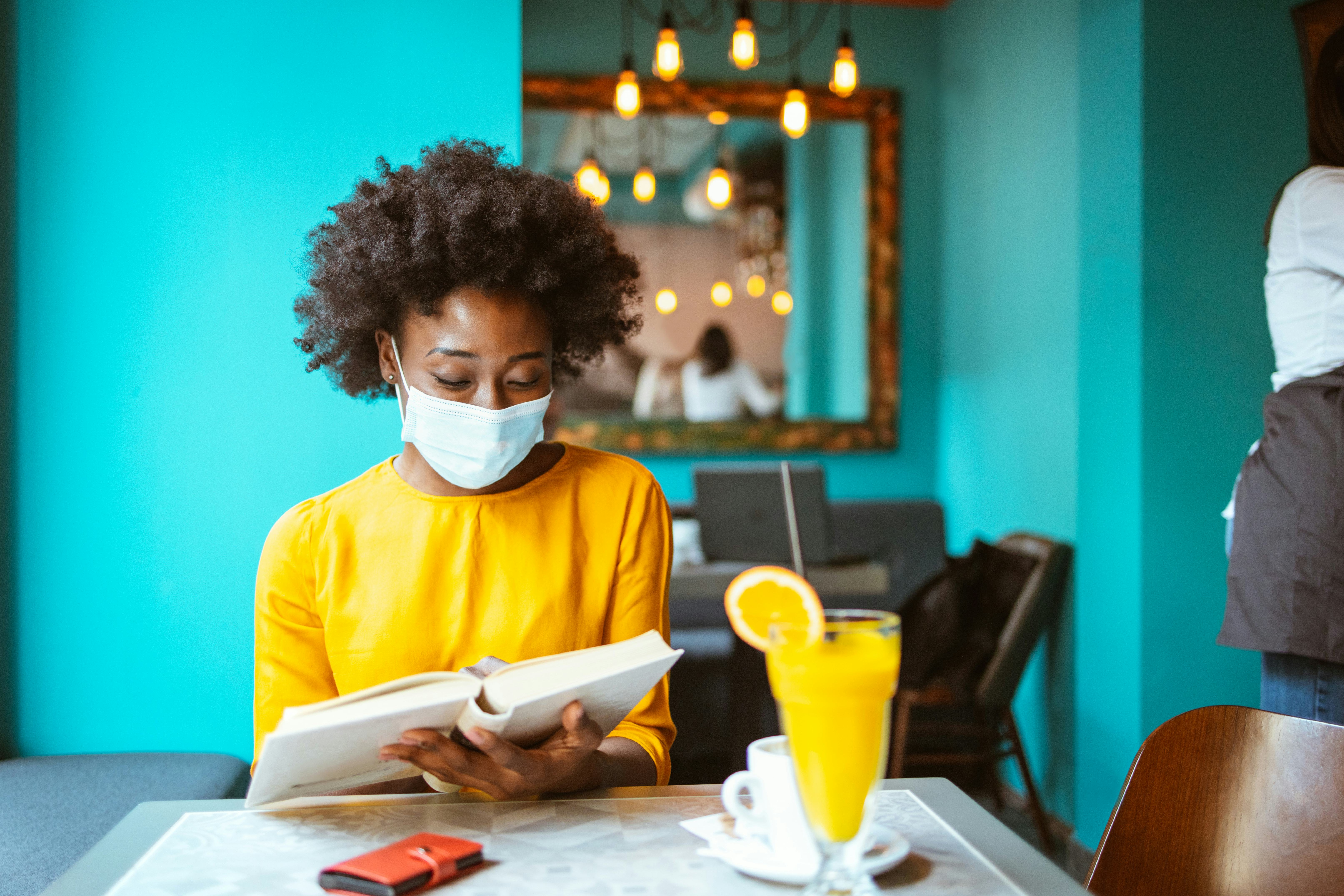 A happy African American woman with protective mask on her face reading book in a coffee shop. New n...