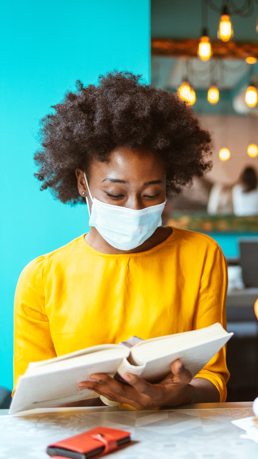 A happy African American woman with protective mask on her face reading book in a coffee shop. New n...