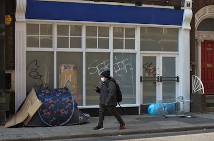 A man wearing a face mask walks past a a rough sleeper's tent outside the entrance to a closed busi...